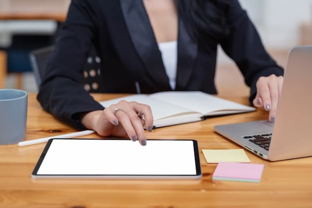 A hand pointing at a digital tablet screen that displays an online unemployment application form. The background shows a person sitting at a desk in a home office, reviewing documents.