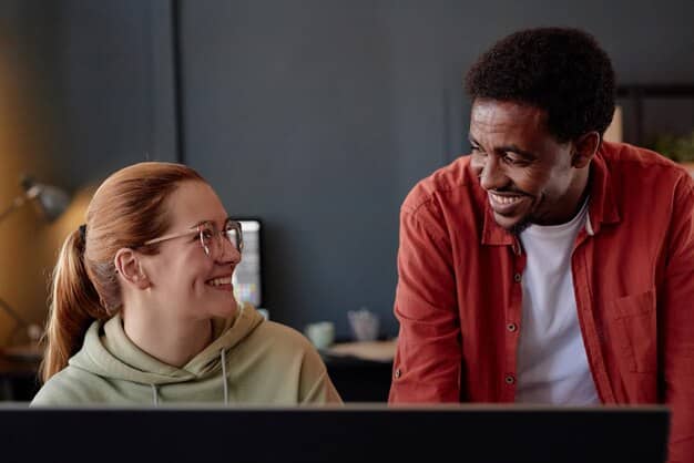 A student smiling confidently while interacting with a mentor in a corporate office environment. Both are looking at a computer screen, suggesting a collaborative project.